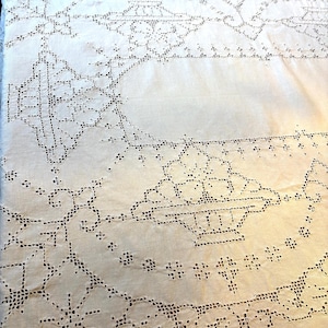May include: Ivory-colored, intricately embroidered tablecloth. The design features geometric patterns, floral motifs, and openwork details. The embroidery is done in a darker shade, creating a contrast against the lighter fabric. The tablecloth is likely made of linen or cotton.