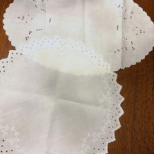May include: Two round white doilies featuring intricate cutwork embroidery and scalloped edges. The top doily displays a delicate floral pattern with eyelet details, while the bottom doily showcases a vine-like stitched border design.