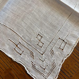 May include: Close-up of a white linen handkerchief with intricate cutwork and a scalloped edge. The detailed design features geometric patterns and delicate floral accents, showcasing a vintage aesthetic. The fabric appears soft and slightly textured.