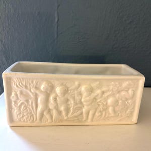 May include: Cream-colored rectangular planter with a relief design of cherubs and foliage. The planter is made of ceramic and has a smooth, glossy finish. The cherubs are depicted in various poses, adding a decorative touch to the piece.