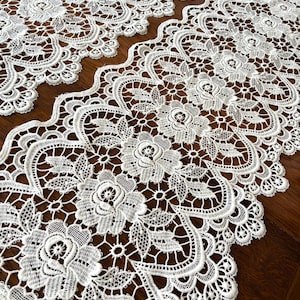 May include: Close-up of white lace fabric with floral and geometric patterns. The lace features detailed rose and leaf designs, with a scalloped edge. The fabric is likely used for decorative purposes, such as table runners or embellishments.