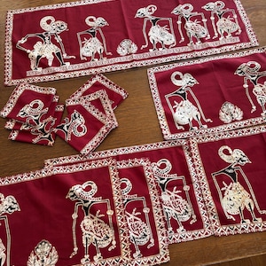 May include: A burgundy table setting featuring a runner, placemats, and coasters. Each piece is adorned with a white and black illustration of a traditional Indonesian puppet figure. The edges are trimmed with a white zigzag pattern.