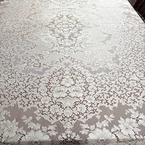 May include: A white lace tablecloth with a floral pattern covers a wooden table. The intricate design features repeating floral and geometric shapes, creating a delicate and elegant aesthetic. The tablecloth is rectangular and appears to be made of a soft, draping fabric.