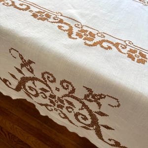 May include: White linen tablecloth with intricate brown cross-stitch embroidery. The design features floral and scroll patterns along the edges and center, creating a vintage aesthetic. The tablecloth is on a wooden surface.