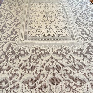 May include: A rectangular off-white lace tablecloth featuring an intricate central panel with swirling patterns, surrounded by a wide border of dense, ornate floral and leaf motifs with a scalloped edge and a fine mesh background.