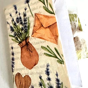 May include: A decorative paper craft featuring a lavender bouquet in a brown sack, a brown envelope, and a wooden heart. Green sprigs and lavender sprigs are also present. The background has text from a book.