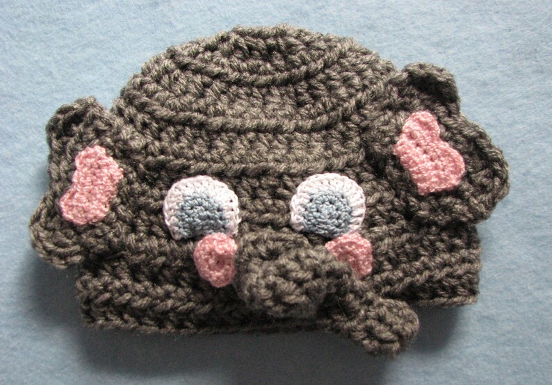 May include: Gray crocheted elephant hat with pink ears, a gray trunk, and blue and white eyes.
