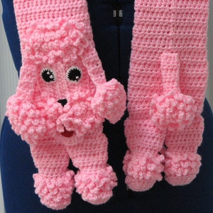 May include: Pink crocheted scarf featuring a poodle design. The poodle has black eyes and a pink nose. The scarf has pom-poms on the ends.
