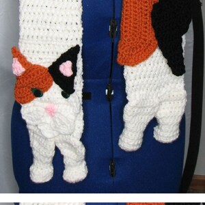 Puede incluir: Una bufanda de crochet blanca, naranja y negra con un diseño de gato. La bufanda se muestra en un maniquí azul.