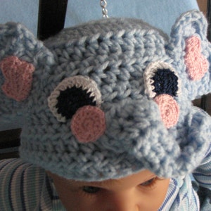May include: A blue crocheted elephant hat with pink and white accents. The hat has a large, floppy ear on each side and a small, pink nose.