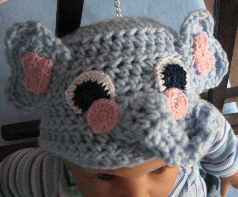 May include: A blue crocheted elephant hat with pink and white accents. The hat has a large, floppy ear on each side and a small, pink nose.