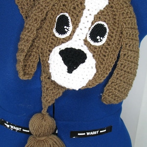 May include: Brown crocheted dog-shaped scarf with a white face and black nose. The scarf has a brown tassel at the end.