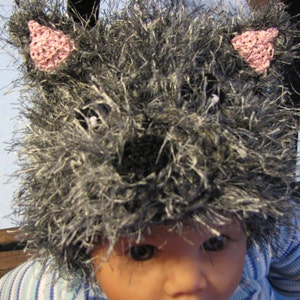 May include: A gray and white fuzzy knit hat with pink ears. The hat is worn by a child with a light complexion and brown eyes.