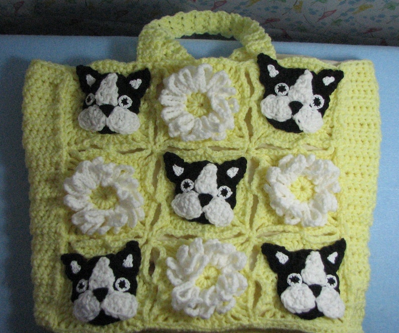 May include: Yellow crocheted tote bag with a square pattern featuring white crocheted flowers and black and white Boston Terrier dog faces.