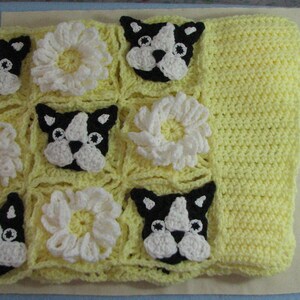 May include: A yellow crocheted baby blanket with a pattern of white flowers and black and white French bulldog faces.