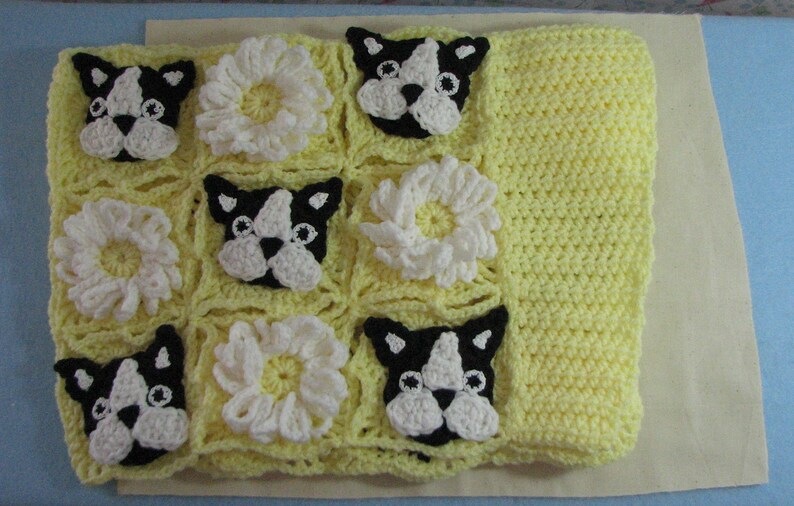 May include: A yellow crocheted baby blanket with a pattern of white flowers and black and white French bulldog faces.