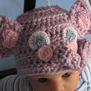 May include: A purple crocheted baby hat with pink, blue, and white crocheted embellishments. The hat is shaped like an elephant with floppy ears.