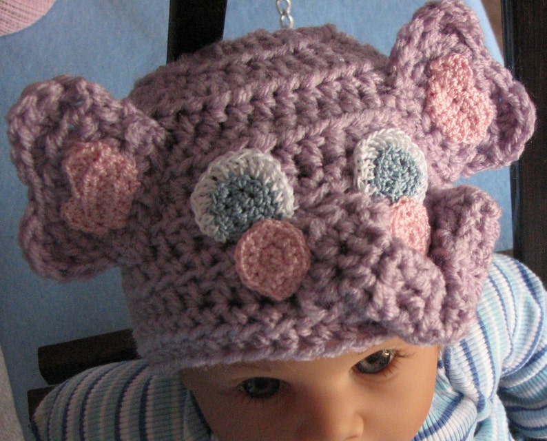 May include: A purple crocheted baby hat with pink, blue, and white crocheted embellishments. The hat is shaped like an elephant with floppy ears.