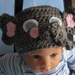 May include: A gray crocheted elephant hat with pink and blue accents. The hat has a long trunk and large ears.
