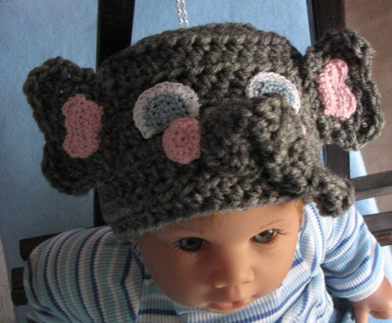 May include: A gray crocheted elephant hat with pink and blue accents. The hat has a long trunk and large ears.