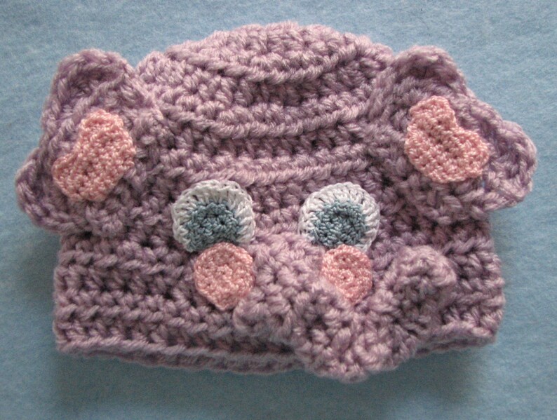 May include: A purple crocheted hat with pink and blue accents. The hat has two large ears and a small nose. The hat is decorated with small crocheted hearts and circles.