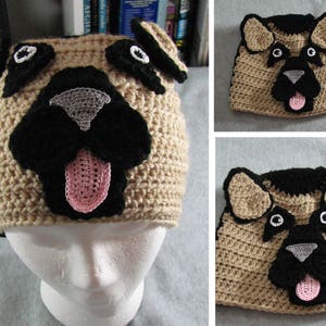 May include: Crocheted dog hat with brown and black fur, black nose, pink tongue, and white eyes. The hat is on a white mannequin head.