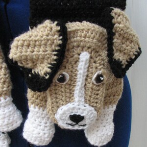 May include: A handmade crochet scarf featuring a dog design. The scarf has a tan body with black and white accents, including the ears, nose, and paws. The dog's face is detailed with black eyes and a white stripe down the nose.