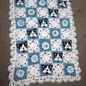 Può includere: Una copertina per neonati in crochet bianco e blu con un bordo a festoni. La copertina presenta un motivo ripetitivo di quadrati blu e bianchi con un piccolo fiore all'uncinetto al centro di ogni quadrato blu. Ci sono anche piccoli cani in crochet bianco e nero al centro di ogni quadrato bianco.