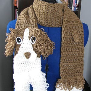 Puede incluir: Una bufanda de ganchillo marrón y blanca con un diseño de cara de perro. El perro tiene pelo marrón, pelo blanco y ojos negros. La bufanda tiene flecos en los extremos.