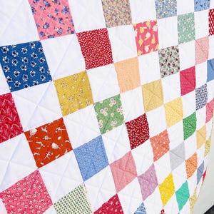 Puede incluir: Un edredón hecho a mano con un diseño de patchwork. El edredón presenta una cuadrícula de cuadrados coloridos con varios patrones, incluyendo estampados florales y geométricos, sobre un fondo blanco. El edredón está acolchado a máquina con un patrón de diamantes.