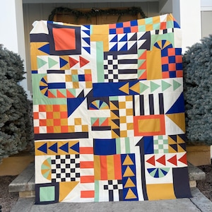 May include: A colorful geometric quilt with a patchwork design. The quilt features squares, triangles, and rectangles in shades of orange, blue, yellow, green, and black. The quilt is displayed outdoors.