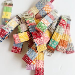 Puede incluir: Una colección de llaveros de tela con un diseño de patchwork y varios patrones coloridos, incluyendo motivos florales, geométricos y de estrellas. Cada correa tiene un cierre de metal plateado en la parte superior para sujeción.