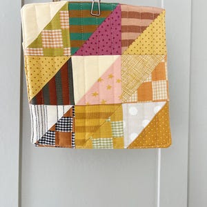 Op de afbeelding: Een vierkante, patchwork gewatteerde pannenlap met een verscheidenheid aan stoffen in tinten geel, groen, roze en bruin. De pannenlap wordt opgehangen met een metalen clip en heeft een kleine lus om op te hangen. Het ontwerp heeft driehoeken en vierkanten.
