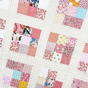 以下が含まれることがあります： パッチワークデザインの手作りキルトのクローズアップ。キルトは、クリーム色の背景に、さまざまなピンク色の花柄と模様の生地の正方形を特徴としています。キルトは、格子模様でミシン縫いされています。