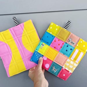 May include: Two colorful square potholders. One is pink and yellow with a curved design. The other is a patchwork of squares in various colors, including blue, pink, and yellow, with star and zebra patterns. Each has a black and white striped loop.