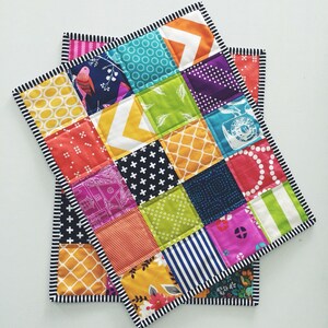 May include: A colourful patchwork quilt with a black and white trim. The quilt is made up of squares of different fabrics, including floral, geometric, and abstract patterns. The quilt is folded in half and is lying on a white surface.