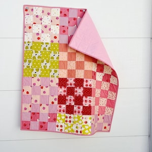 Könnte beinhalten: Ein Patchwork-Quilt mit rosa Rand und Rückseite. Die Vorderseite zeigt Quadrate in Rosa, Rot und Grün mit Erdbeer- und Apfelmustern. Der Quilt hängt an einer weißen Wand.