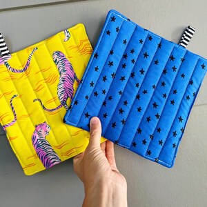 May include: Two square quilted pot holders with hanging loops. One is bright yellow with a pink and black tiger pattern. The other is vibrant blue with a black star print. Both feature vertical stitching and black and white striped fabric loops.