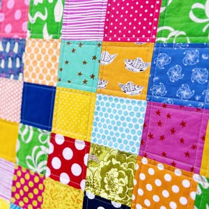 May include: A colorful patchwork quilt with a variety of patterned squares. The squares feature polka dots, stars, floral designs, and other playful patterns in shades of red, pink, green, blue, yellow, and orange. The quilt is machine-stitched.