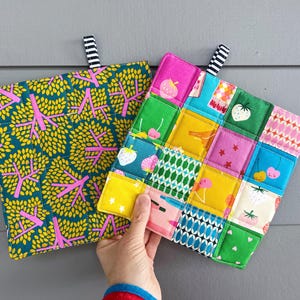 May include: Two colorful quilted pot holders. One features a pattern of yellow leaves and pink tree branches on a teal background. The other is a patchwork design with fruit and floral patterns in various colors. Each has a black and white striped loop.