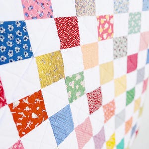 May include: A patchwork quilt with a grid pattern. The quilt features squares of white fabric interspersed with colorful patterned squares in shades of pink, blue, red, yellow, and green. The quilt is likely handmade.