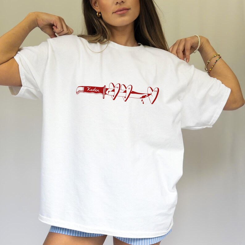 May include: White t-shirt with a red graphic design. The design features a knife with the word "Xaden" and several heart shapes. The hearts have a dripping blood effect. The shirt is a casual, oversized style.