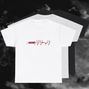 May include: White t-shirt featuring a red knife and heart graphic, with the word "Xaden" on the knife. The image shows three t-shirts in white, grey, and black, layered together.