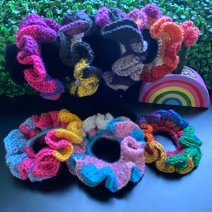 May include: A set of colorful crocheted hair scrunchies in various colors, including pink, purple, blue, yellow, orange, and green. The scrunchies are arranged in a circle on a dark blue surface.