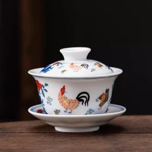 May include: White ceramic teacup and saucer set with a rooster and chick design. The cup has a lid and the saucer has a blue rim.