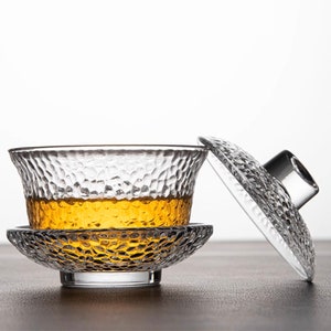 May include: A clear glass teacup with a textured surface and a matching lid. The teacup is filled with a light amber tea.