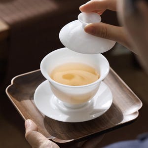 May include: A white ceramic teacup and saucer with a lid, filled with light brown tea, sitting on a dark brown wooden tray.