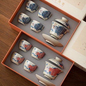 Könnte beinhalten: Zwei Sets chinesischer Teetassen und Teekanne in Geschenkboxen. Die Teetassen und die Teekanne sind mit einem blauen und weißen Fischdesign verziert. Die Boxen sind mit "Jin Yu Man Tang" beschriftet.