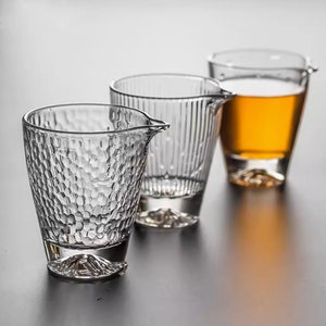 Könnte beinhalten: Drei klare Glas-Teetassen mit einem strukturierten Design. Die Teetassen haben einen kleinen Ausguss und einen abgerundeten Boden. Die Teetasse rechts enthält eine hellbraune Flüssigkeit.