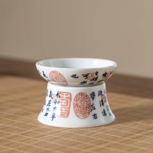 Op de afbeelding: Een witte theekop van keramiek met blauwe en rode Chinese karakters staat op een bijpassende witte keramische standaard. De theekop en standaard zijn beide versierd met een herhalend patroon van Chinese karakters.
